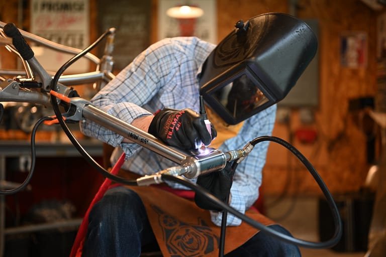 A framebuilder in a welding mask leans over to apply a torch to a bicycle frame under construction.
